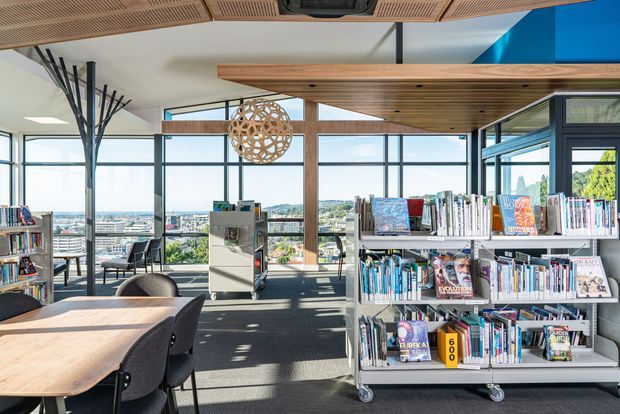 A bright contemporary library or reading area with extensive floor-to-ceiling glazing, rolling bookshelves, timber-framed ceiling features, reading tables and chairs, and a panoramic urban view. The space has a refined, warm modern fit-out with strong natural light and open-plan planning.