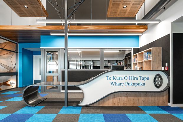Contemporary commercial interior reception or entry space featuring a sculptural curved reception desk with bilingual text, timber-faced built-in shelving, vivid blue and charcoal wall accents, patterned carpet tiles, exposed structural elements, and timber acoustic ceiling panels.