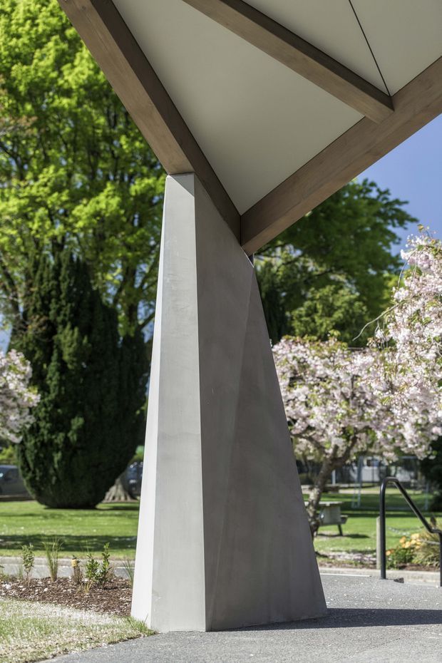 Exterior architectural detail of a contemporary pavilion or canopy in a landscaped park-like setting. A sharply angled, tapered concrete or rendered support rises to meet a broad projecting roof with exposed dark timber beams and a smooth light soffit. Mature trees, flowering vegetation, lawn, and paved paths surround the structure.