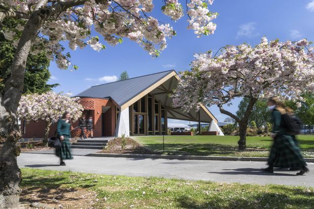 Exterior view of a contemporary gable-roofed building set within a landscaped campus or public garden. The building features a steep metal roof, glazed front facade, exposed timber framing, and a contrasting red brick side volume. Flowering trees, lawn, paved walkways, and blurred pedestrians surround the structure.