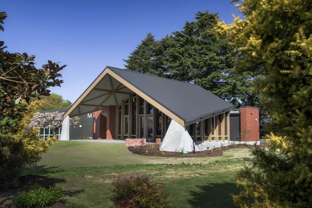 Exterior view of a contemporary pavilion-like building set within a landscaped garden. The structure has a prominent steep gabled roof with dark metal roofing, broad glazed façades, exposed timber framing beneath the gable, dark horizontal wall cladding, and a contrasting red brick chimney or feature wall. Mature trees and lawn surround the building.