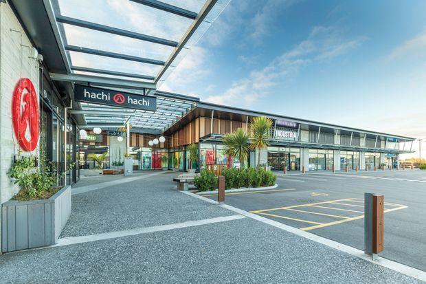 Exterior view of a contemporary single-level commercial retail and dining complex, featuring a broad glazed canopy over the pedestrian forecourt, dark metal framing, timber-toned facade cladding, extensive shopfront glazing, landscaped raised planters, and marked parking or loading areas. Visible signage includes hachi hachi and other retail tenants. The image is captured in bright daylight from the open forecourt.