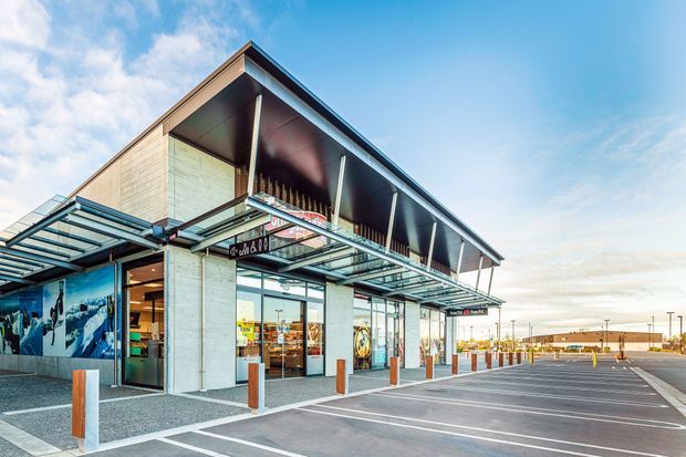 Exterior view of a contemporary low-rise commercial retail building with a prominent deep overhanging roof, exposed slim columns, extensive glass storefronts, and a covered glazed canopy along the entrance. The building sits beside a large marked asphalt parking area with bollards and pedestrian paving. Storefront signage and merchandise are visible, but the primary subject is the building exterior.
