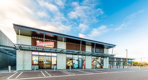 Exterior view of a contemporary single-storey commercial retail building with a broad projecting roof, exposed structural elements, concrete masonry walls, extensive storefront glazing, and colourful shopfront signage. The image shows a paved parking and pedestrian forecourt under a bright sky.