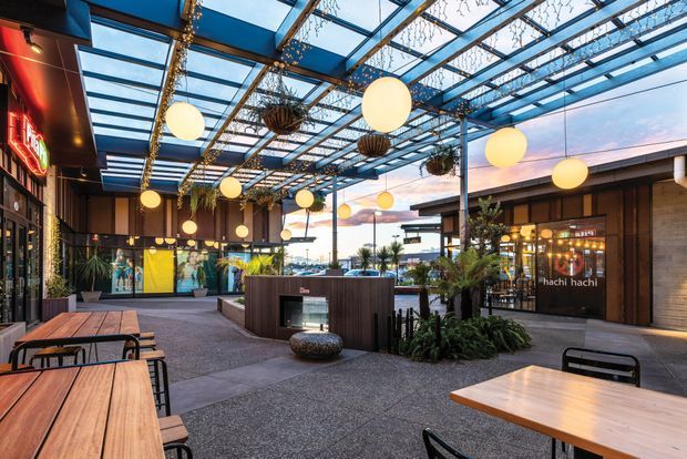 A covered outdoor dining and hospitality precinct with timber tables, black metal chairs, planted garden beds, and a central service or planter enclosure. The space is enclosed by a prominent blue-painted steel pergola with transparent roof panels, decorated with hanging greenery and numerous warm globe lights. Restaurant and retail frontages line the edges, with a visible “hachi hachi” storefront. The twilight sky and open-air arrangement create a lively, contemporary urban dining atmosphere.