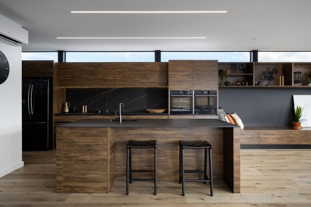 Contemporary open-plan kitchen featuring a large dark timber island with integrated sink and seating for two, matching timber cabinetry, black stone-look splashback, black appliances, and open display shelving. The room has a light timber floor, dark grey wall finishes, slim linear ceiling lights, and a long horizontal clerestory window.