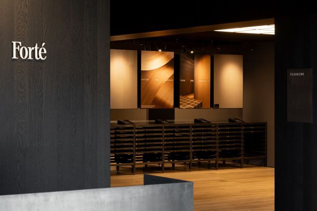 A dark, refined flooring showroom interior with warm timber flooring, black display shelving, wood product sample panels, and dramatic integrated lighting. A large dark timber-textured wall bears the “Forté” brand, while a concrete-toned reception counter occupies the foreground.