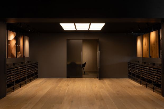 A dark, sophisticated interior entry or reception space with warm timber flooring, deep brown walls and ceiling, integrated low storage or display cabinetry lining both sides, and illuminated timber feature panels. A central doorway opens to a smaller room beyond, while a rectangular ceiling light provides a dramatic focal point.