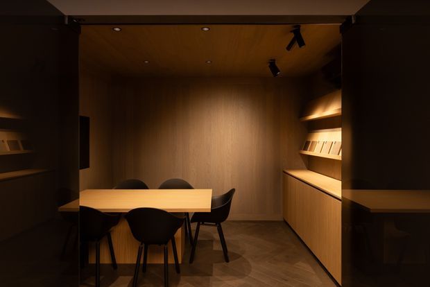A compact, dark-toned wood-lined interior resembling a private dining room or meeting room. A timber table with several black chairs occupies the centre, while built-in timber cabinetry and illuminated open shelving line the right wall. Recessed downlights and black track spotlights provide warm, focused lighting, and the floor has a dark herringbone timber finish.