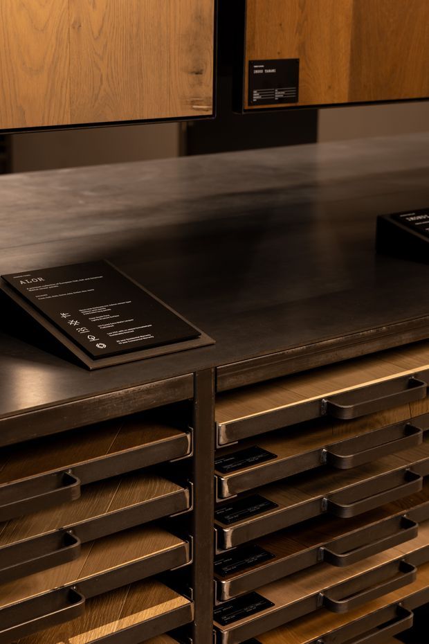 Close-up view of a dark timber display cabinet or merchandising console with multiple shallow drawers, bronze-toned metal drawer fronts and handles, and a broad dark countertop. Timber sample panels and black informational plaques are displayed above and on the surface, suggesting a material or interior finishes showroom. The scene is warmly lit with a refined, dark-toned presentation.