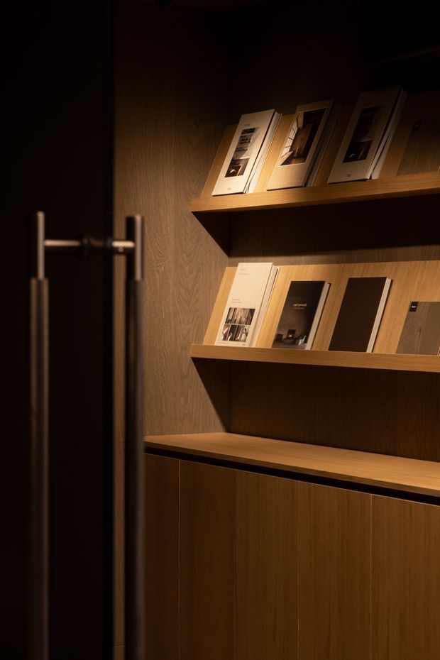 A dimly lit, refined built-in display niche viewed through a partially open dark door. Warm timber-veneer wall panelling, floating shelves, and lower closed cabinetry display books, brochures, and material samples, creating an intimate showroom or entry display area. A tall brushed-metal door pull is visible in the foreground.