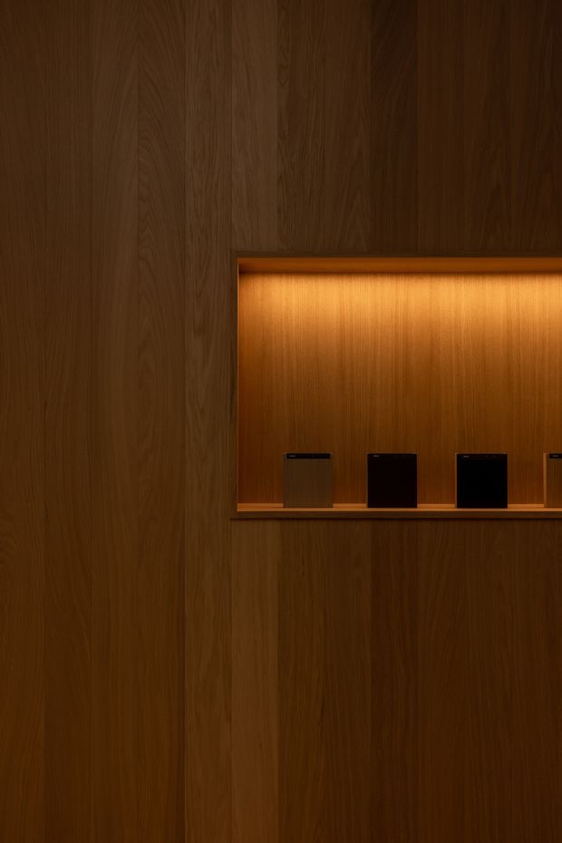 A dark, refined timber-lined interior wall featuring a recessed illuminated display niche with several small dark objects arranged on a shelf. Warm integrated lighting highlights the rich vertical wood grain and creates an intimate, gallery-like atmosphere.