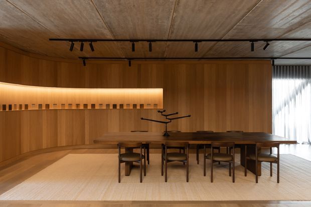A warm, wood-lined dining room with a long rectangular dining table and multiple dark wood chairs. The room features full-height timber wall and ceiling panelling, a softly illuminated recessed wall display or shelf, minimalist black track lighting, and sheer curtains covering a large window on the right. A sculptural black centerpiece sits on the table, creating a refined, intimate atmosphere.