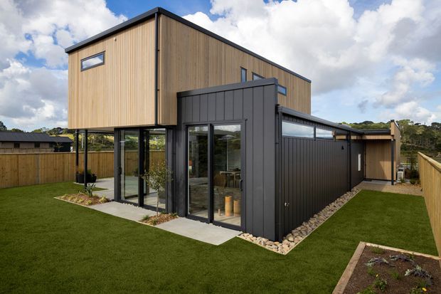 Exterior view of a contemporary two-storey home with a box-like upper volume clad in warm vertical timber, a dark grey metal-clad lower wing, extensive floor-to-ceiling glazing, and a flat roof. The house sits within a fenced landscaped yard with lawn, garden beds, and a gravel border.