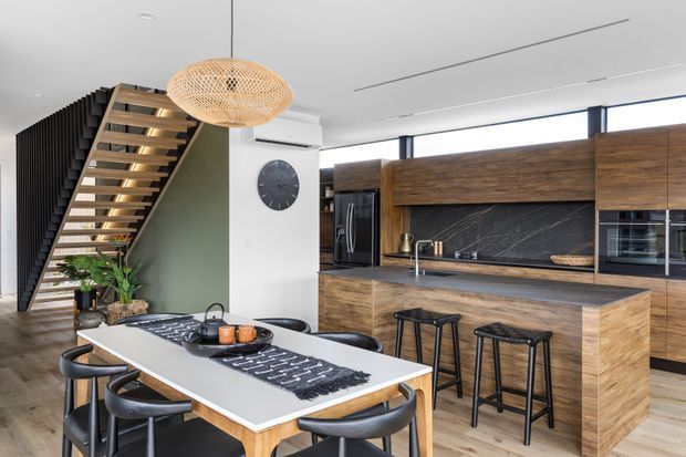 A contemporary open-plan kitchen and dining area with extensive dark timber cabinetry, a black marble-look splashback, a large white island with seating, and a timber dining table. A sculptural timber staircase with black vertical screening rises beside the kitchen, while a woven pendant light and indoor plants add warmth.