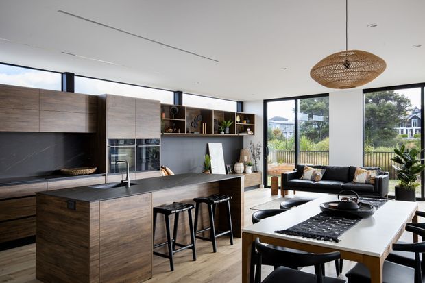 A bright, open-plan contemporary kitchen and living/dining area with extensive floor-to-ceiling glazing. The kitchen features dark wood cabinetry, a large island with a dark benchtop and integrated sink, open shelving, and black appliances. A black sofa and dining table occupy the adjoining living and dining zones. A woven pendant light, indoor plants, and views to a landscaped residential setting add warmth and texture.