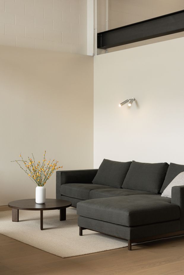 A minimalist living room with a dark charcoal L-shaped sectional sofa and ottoman, a low round dark coffee table, and a simple white vase with delicate branches. The room has warm pale walls, light timber flooring, a neutral rug, a wall-mounted spotlight, and a high ceiling with an exposed dark steel beam and upper blockwork detail.