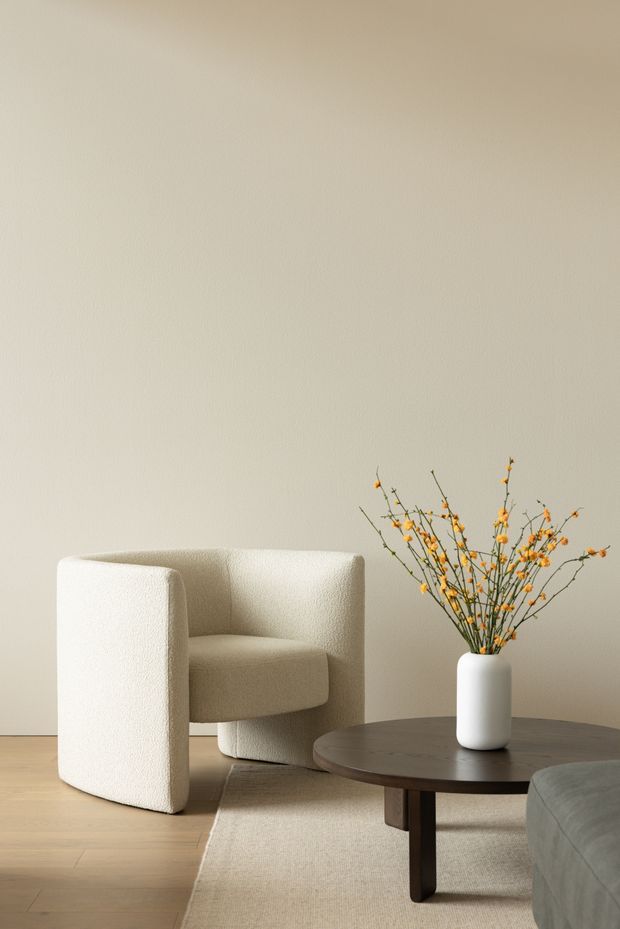 A softly lit minimalist living room vignette featuring a sculptural cream boucle armchair, a dark round wooden coffee table, a white ceramic vase with delicate orange branches, pale timber flooring, and a neutral textured rug. The warm off-white wall and restrained furnishings create a calm, refined atmosphere.