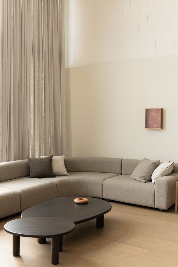 A calm, softly lit living room featuring a large curved taupe sectional sofa, layered neutral cushions, dark rounded coffee tables, full-height beige curtains, pale timber flooring, and a minimalist framed artwork. A high wall or ceiling bulkhead with subtle rectangular panel lines adds architectural interest.
