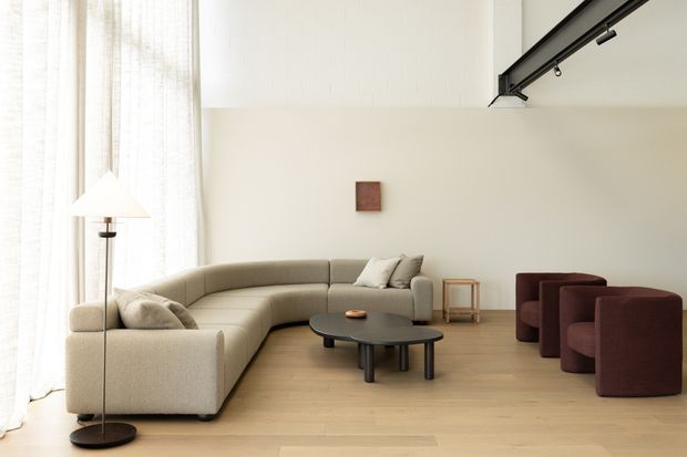 Bright, spacious contemporary living room with a large curved beige sectional sofa, low black oval coffee table, burgundy lounge chairs, pale timber flooring, sheer floor-to-ceiling curtains, and a slim black ceiling-mounted track light. The room has a restrained, minimalist composition with high ceilings and abundant natural light.