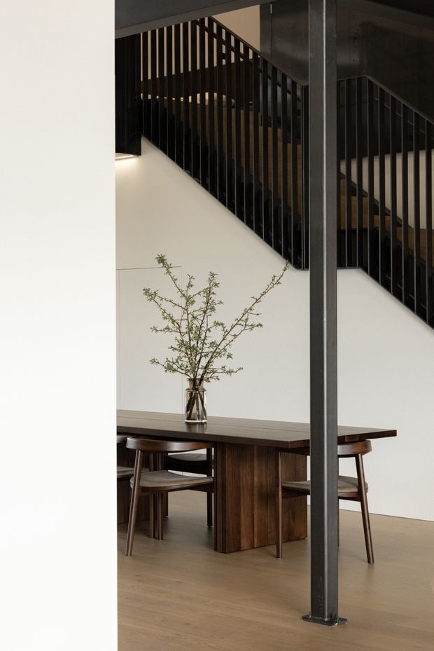 A refined interior view showing a dark timber dining table and chairs beneath a staircase. The staircase features slim black metal balustrades and a substantial dark structural post, contrasted against white walls and light timber flooring. The space has a restrained, contemporary minimalist character with warm wood tones and integrated stair lighting.
