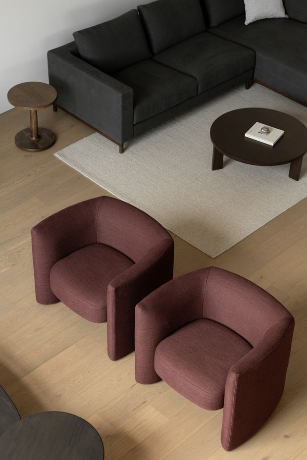 A contemporary living room photographed from above, featuring a dark charcoal sectional sofa, two burgundy upholstered tub chairs, a pale textured area rug, light timber flooring, and dark round coffee and side tables. The restrained palette and clean furniture forms create a refined, modern seating area.