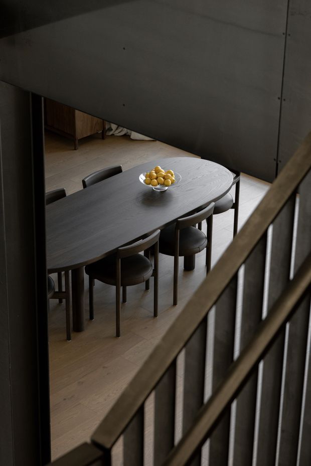 A dark, refined dining area viewed from an upper stair or landing. A large organically shaped dark timber dining table is surrounded by upholstered dining chairs, set over pale timber flooring. A simple bowl of yellow fruit provides the only strong colour accent. The space is partially screened by a substantial dark grey wall or ceiling plane and a timber-and-dark-metal stair balustrade in the foreground, creating a moody architectural composition.