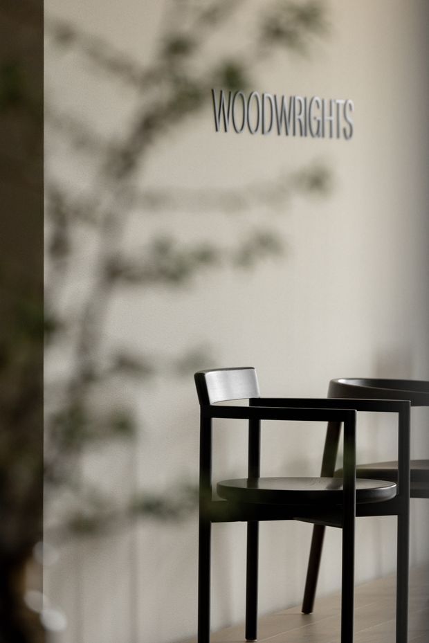 A minimalist dining area with a black rectangular table and matching dark wooden chairs against a warm off-white wall. Soft, blurred plant shadows fall across the wall, and the word “WOODWRIGHTS” is displayed near the upper centre. The scene has subdued lighting and a refined, quiet atmosphere.