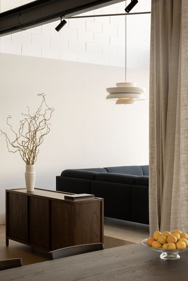 A refined open-plan living and dining interior with a dark modular sofa, walnut-toned sideboard, timber dining table, sculptural layered pendant light, and linen curtains. White-painted masonry walls and exposed dark ceiling beams create a calm, gallery-like setting, with a minimalist vase of branches and a bowl of fruit adding organic detail.