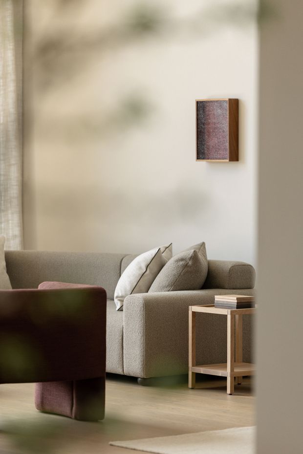 A softly lit, refined living room vignette featuring a low-profile taupe upholstered sofa with neutral cushions, a deep burgundy and green accent armchair, a small timber side table with books, pale timber flooring, a light neutral wall, and a small framed abstract artwork. The room has a calm, warm, contemporary minimalist character with subtle tonal layering and gentle natural light.