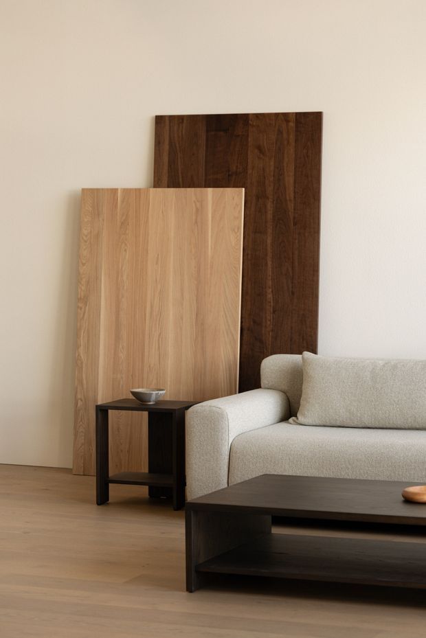 A calm, minimalist living room vignette with a cream upholstered sofa, dark timber coffee table and side table, and two large vertical timber panels leaning against a warm off-white wall. The room has a light timber floor and a restrained natural palette with strong light-and-dark wood contrast.