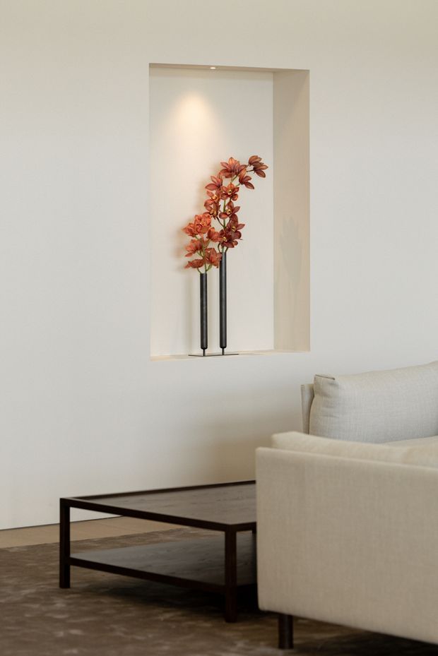 A calm, minimalist living room vignette with a cream upholstered sofa, dark rectangular coffee table, and neutral textured rug. A recessed wall niche with subtle downlighting displays two tall dark vessels holding sculptural reddish-orange floral branches. The space has smooth off-white walls, restrained furnishings, and soft natural lighting.