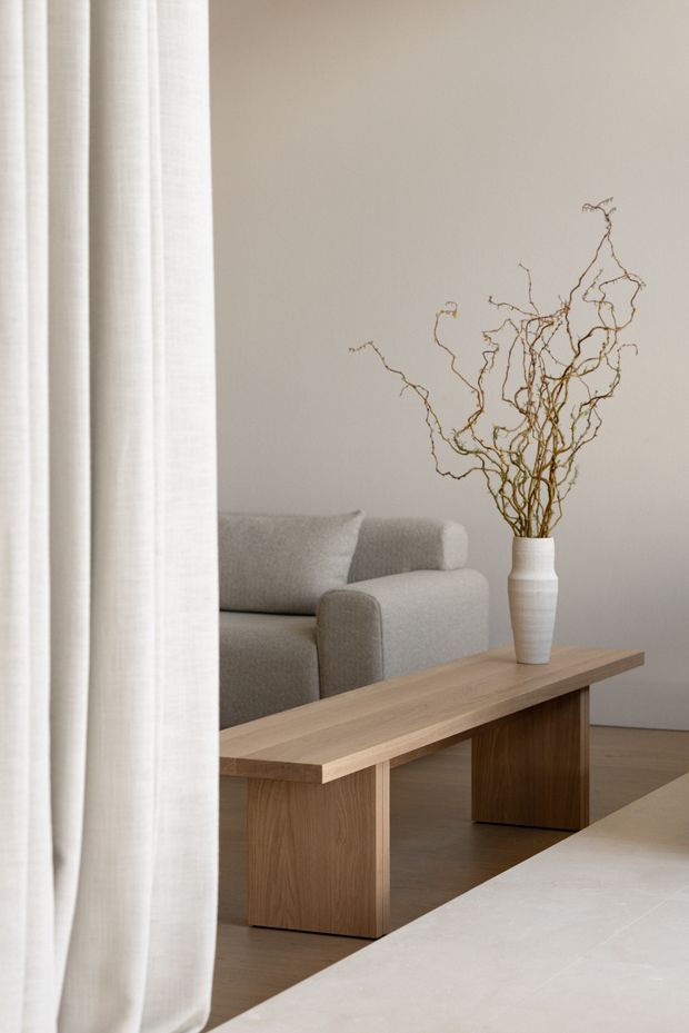 A softly lit minimalist living room viewed past sheer white curtains. The room features a pale upholstered sofa, a simple light timber coffee table with block legs, a white sculptural vase filled with bare branches, warm timber flooring, and neutral plaster walls. A light stone or pale surface is visible in the foreground.
