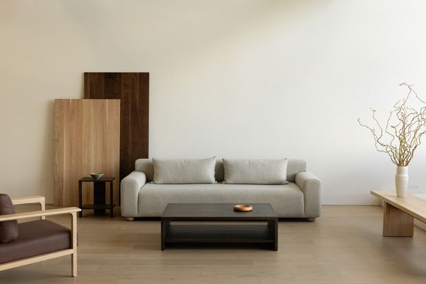 Minimalist living room with a low-profile light grey sofa, dark rectangular coffee table, pale timber flooring, and a simple timber console on the right. Two large timber panels lean against the wall behind the sofa, while a vase of bare branches adds a natural accent. The room is softly lit with a restrained neutral palette and clean, uncluttered composition.