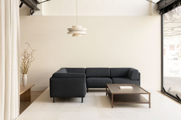 Bright, minimalist living room with a dark charcoal sectional sofa, low rectangular wood-and-metal coffee table, pale tiled floor, warm off-white walls, and a sculptural white pendant light. Black exposed ceiling track elements and large glazing add a subtle contemporary-industrial character. A narrow timber console with a simple vase and branches sits to the left.