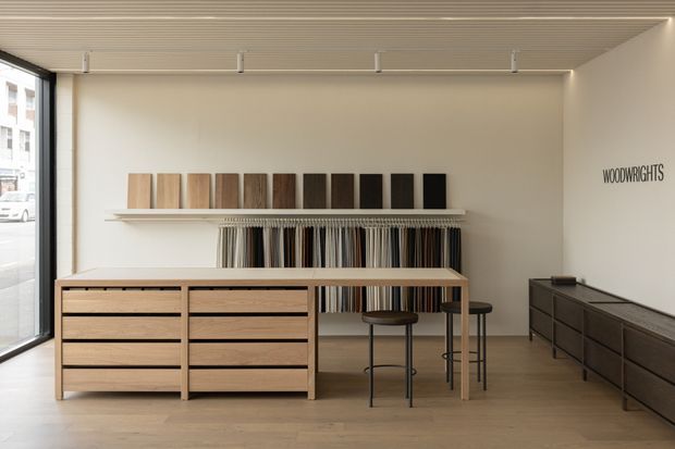 A minimalist woodworking showroom interior with pale timber flooring, a white wall, streamlined display shelving, timber sample boards, hanging material samples, low timber drawer units, and simple black stools. Large glazing on the left provides street views, while the wall is marked with the word “WOODWRIGHTS.”