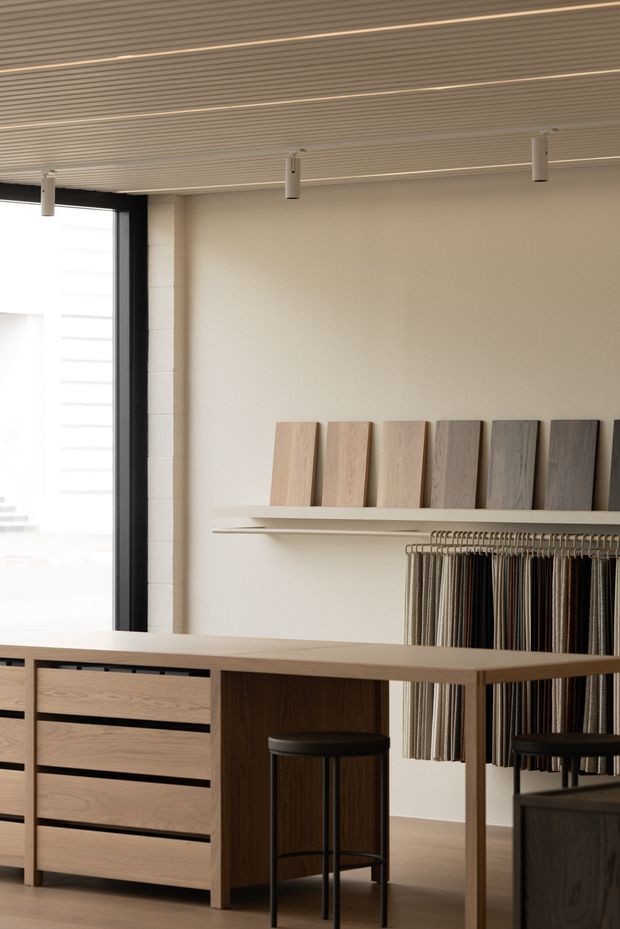 A refined material and flooring showroom interior with a long timber-toned display counter, integrated drawers, black stools, wall-mounted wood sample boards, and hanging textile or flooring samples. The space has warm off-white walls, a ribbed linear ceiling with recessed lighting, dark-framed glazing, and a clean, minimal presentation.
