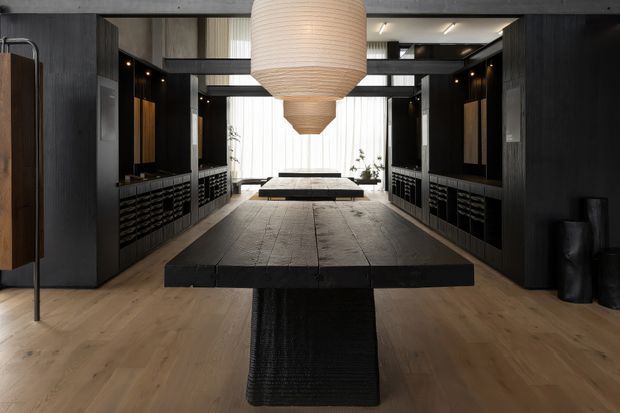 A dark, refined contemporary interior dominated by a long, substantial black dining table with a textured pedestal base. Matching black built-in cabinetry and display shelving line both sides of the room, while warm timber flooring and dark structural beams create strong contrast. Oversized cream paper pendant lights hang along the central axis, with sheer curtains and a secondary table visible at the far end.