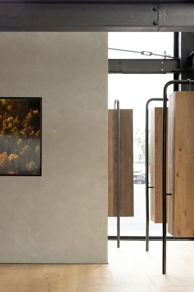 A minimalist interior entry area featuring a smooth warm-grey plaster wall, pale timber flooring, a framed artwork, and a large glazed opening with exposed dark steel structural framing. Tall timber pivot or swing doors with slender black metal handles create a strong industrial-modern threshold, with daylight and a vehicle faintly visible outside.