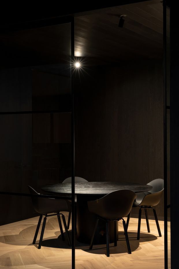 A dark, moody dining area viewed through black-framed glass partitions. A round sculptural dining table with several curved dark chairs sits beneath a timber-lined ceiling and a focused pendant or spotlight. The space features rich dark timber wall panelling and warm herringbone timber flooring, with dramatic low-level lighting and strong shadows.