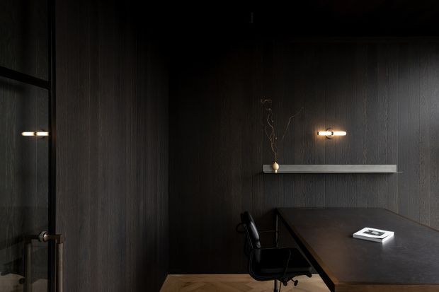A dark, refined home office with full-height charcoal vertical wall panelling, a large dark wood desk, black office chair, minimalist wall shelf, and warm linear wall sconces. A glass door or partition is visible on the left, with subtle timber flooring below. The atmosphere is intimate, moody, and sophisticated.