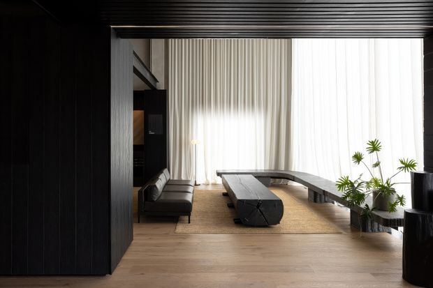 A refined contemporary living room viewed through a dark timber-framed opening. The space features light timber flooring, a large neutral rug, a low black sectional sofa, a sculptural dark coffee table, and a long curved bench beneath full-height sheer curtains. Vertical ribbed wall and ceiling detailing, dark built-in joinery, and potted greenery create a dramatic yet calm interior.