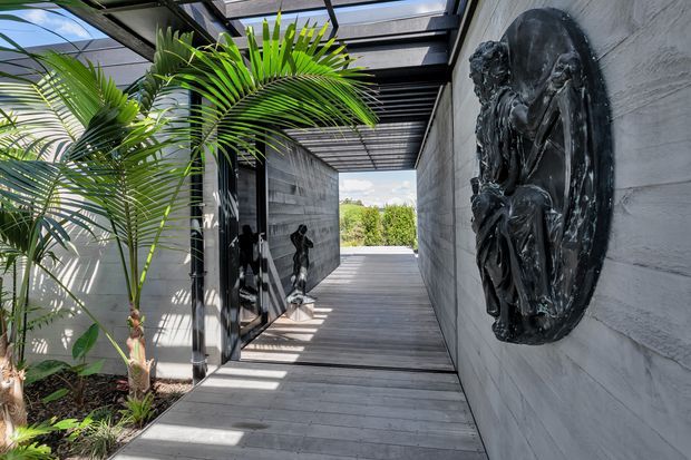 A covered outdoor passageway with grey timber decking, concrete or rendered walls, black structural framing, and a translucent roof. Tropical palms line one side, while dark sculptural wall art and a smaller statue accent the corridor. The passage opens toward a green landscape and bright sky beyond.