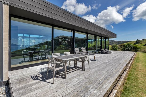Exterior view of a low, contemporary home with a broad flat roof, dark horizontal cladding, extensive floor-to-ceiling glazing, and a large weathered timber deck furnished with an outdoor dining table and chairs. Rolling green landscape is visible beyond the house under a bright blue sky.
