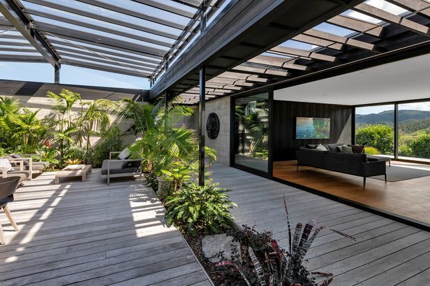 A contemporary indoor-outdoor living area opening onto a spacious timber deck courtyard. Black steel framing supports a translucent and slatted overhead pergola, while lush tropical planting borders the deck. Large glazed openings connect the deck to a dark-clad living room with a sofa, television, and views toward a green hillside.