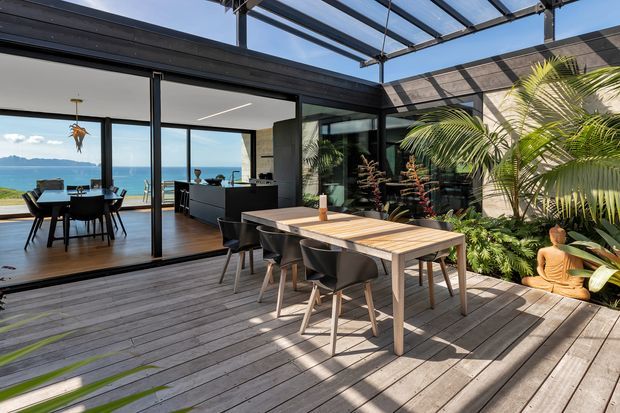 A bright contemporary outdoor dining deck beneath a glazed overhead roof, opening into a modern kitchen and dining area. A large timber dining table with dark chairs sits on weathered timber decking, surrounded by lush tropical planting. Full-height glazing reveals a blue coastal landscape beyond.