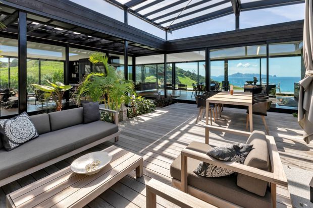 A bright, open-plan indoor-outdoor living and dining space with extensive black-framed glazing, a transparent roof structure, timber decking, and contemporary timber furniture. The room opens toward a vivid coastal outlook with blue ocean and mountainous terrain visible beyond the glazing. Potted palms and plants soften the modern architectural setting.