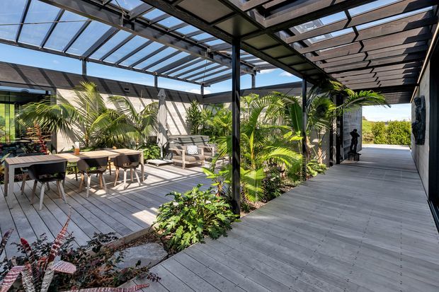 A bright, expansive outdoor entertaining area with a timber deck, dining table and chairs, lounge seating, and abundant tropical planting. A dark metal-framed pergola with translucent roof panels and slatted overhead sections creates covered outdoor zones, while a wide deck walkway leads toward the garden and distant water or sky beyond.