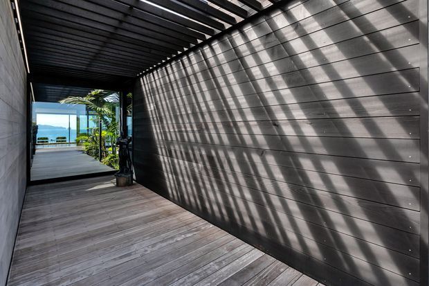A covered exterior walkway or deck corridor with dark horizontal timber wall cladding, matching overhead slatted screening, and grey timber decking. Strong linear shadows fall across the wall and floor. The passage opens toward a planted outdoor area with a distant water view.