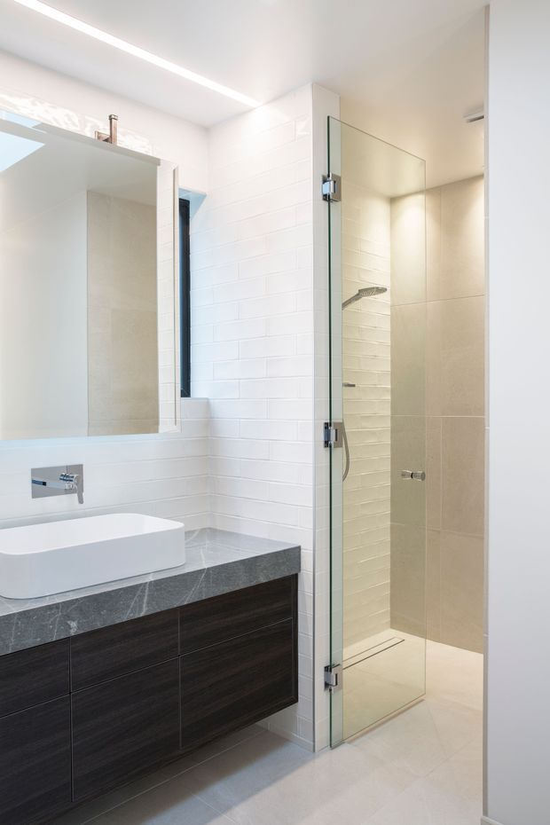 Bright contemporary bathroom with a white vessel basin on a dark wood floating vanity, grey stone benchtop, large mirror, white subway-tile wall, and a frameless glass walk-in shower with recessed shelving and pale floor tiles. Clean-lined integrated lighting and chrome fixtures create a crisp, modern finish.